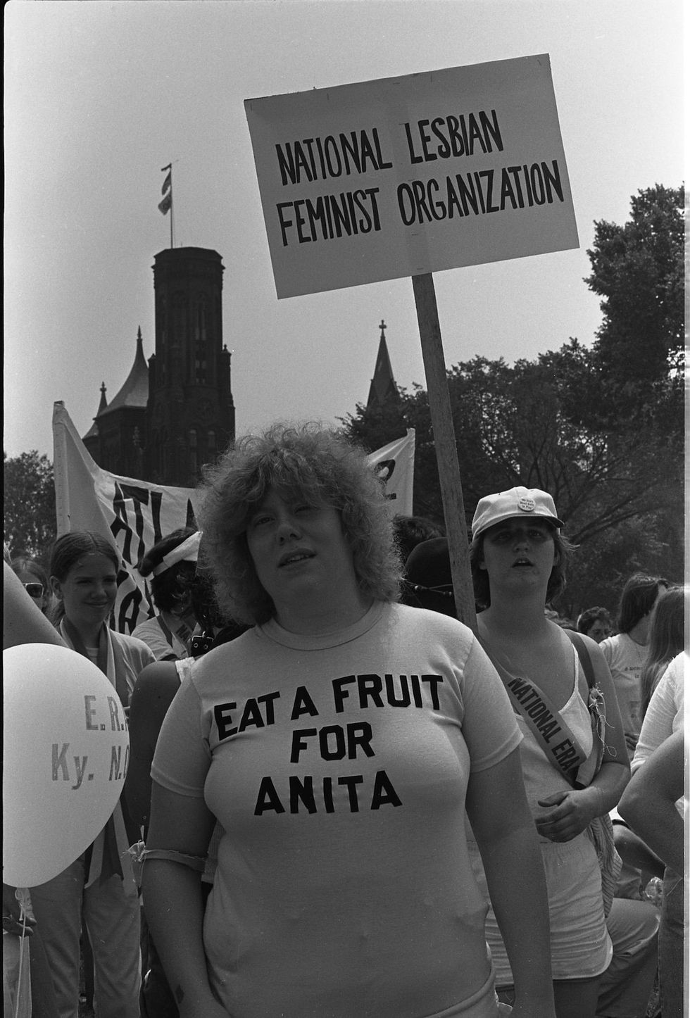 Portrait of a woman standing under the National Lesbian Feminist Organization sign on the National Mall before the Equal Rights Amendment March, Washington, DC, July 9, 1978. She is wearing a T-shirt that says ``Let's eat fruit for Anita.'' (An ironic reference to American singer and Florida Citrus spokesperson Anita Bryant, who is a vocal opponent of laws enacted to prevent discrimination based on sexual orientation). The Smithsonian's towers (also known as castles) are visible in the background.