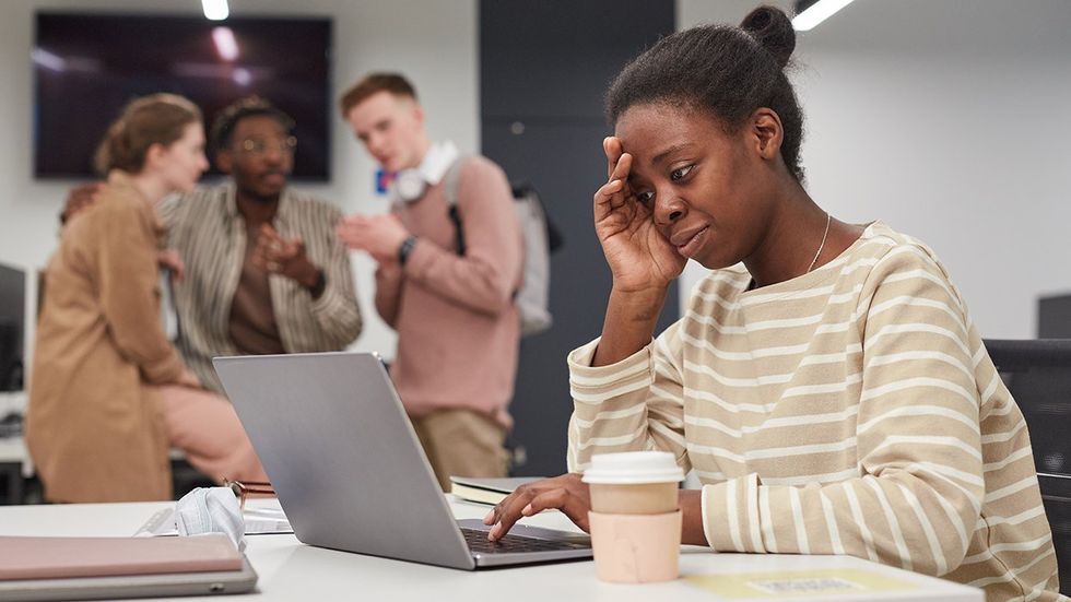 Side view portrait of young African-American woman using laptop alone at table with group of people whispering and bullying her in background